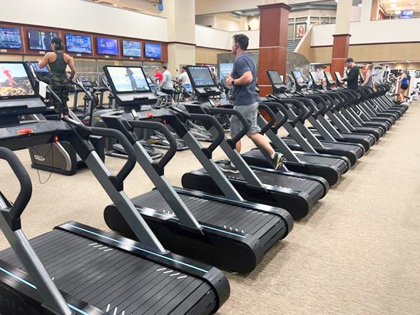 Several people are exercising on treadmills in a spacious gym, with TV screens mounted in front of the machines and gym equipment visible in the background.
