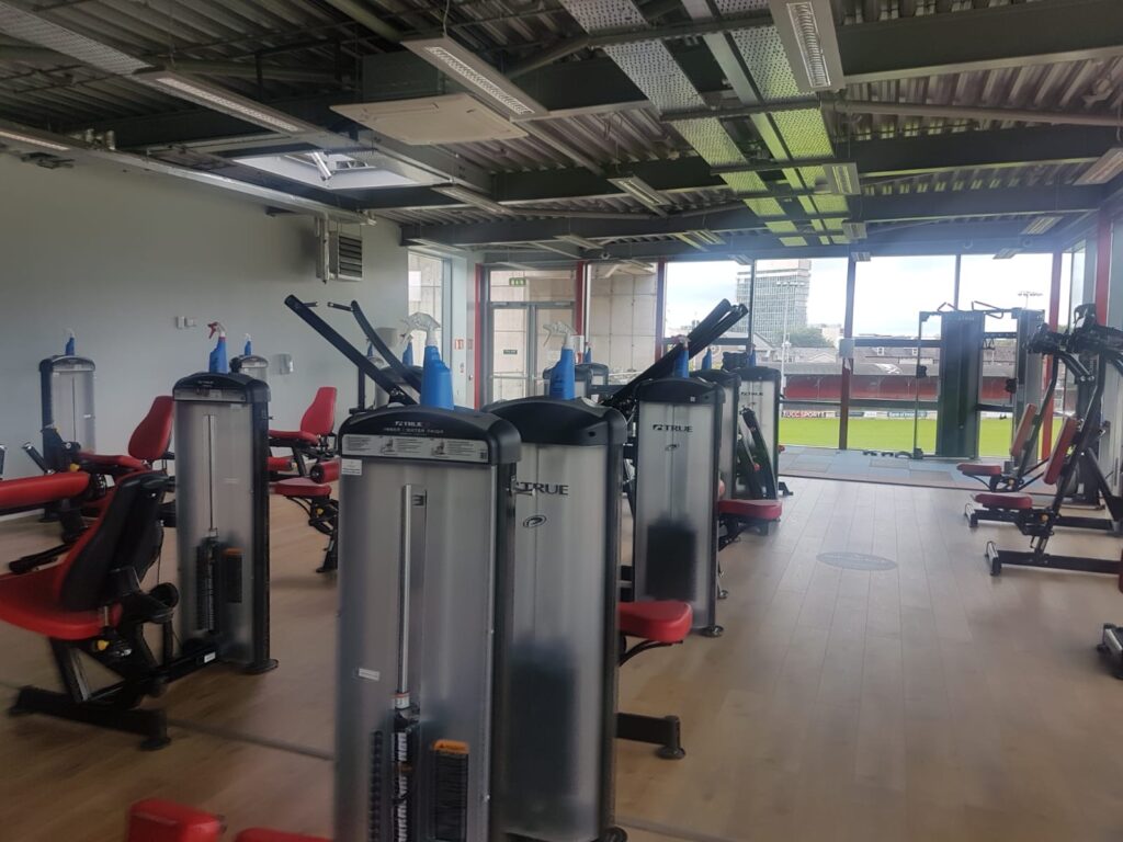 A modern gym with various strength training machines, red and black seats, and large windows offering a view of a sports field and city buildings outside. The gym is well-lit with natural and artificial light.