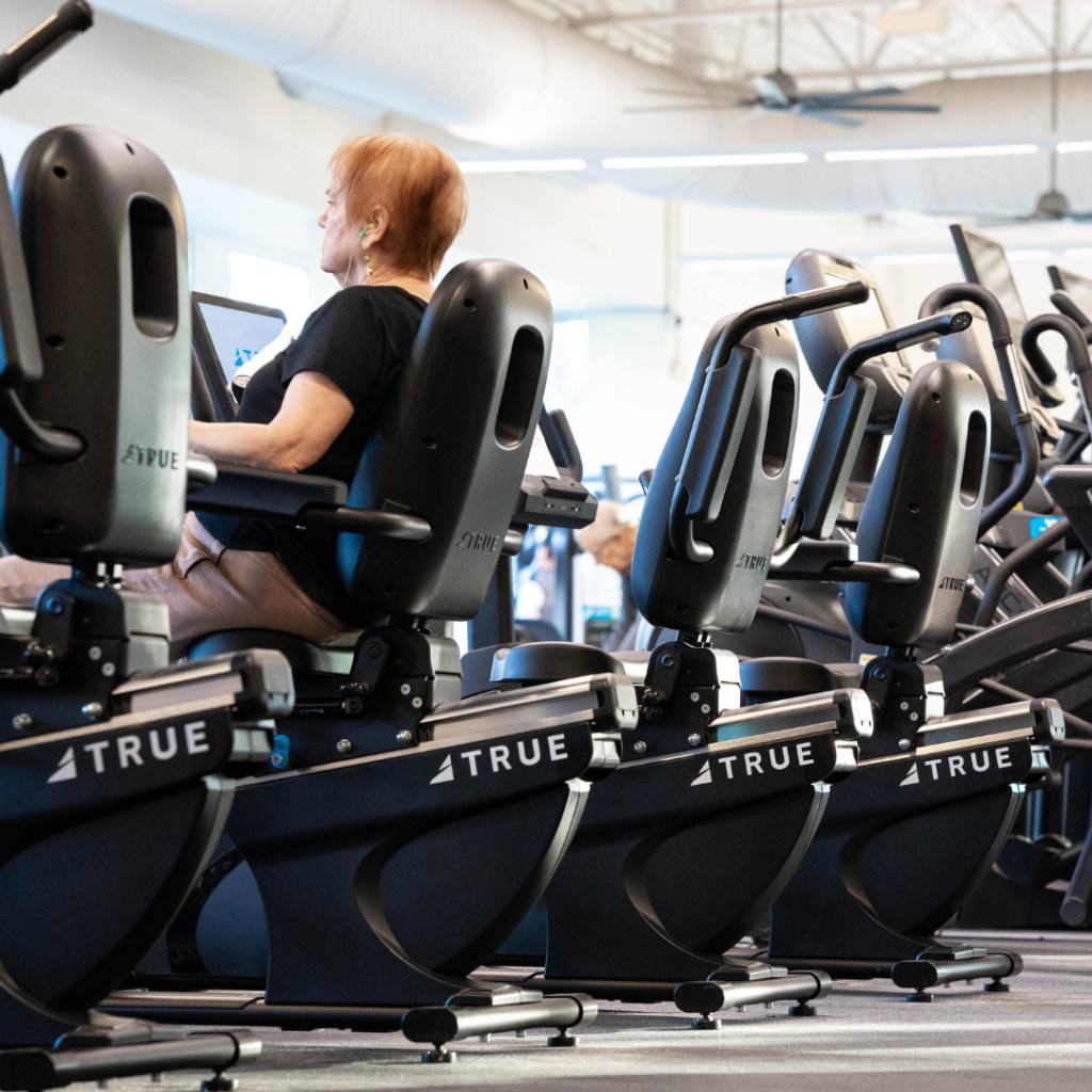 An older woman exercises on a recumbent bike in a gym, sitting among a row of similar machines. The gym is well-lit with large windows and a high ceiling.