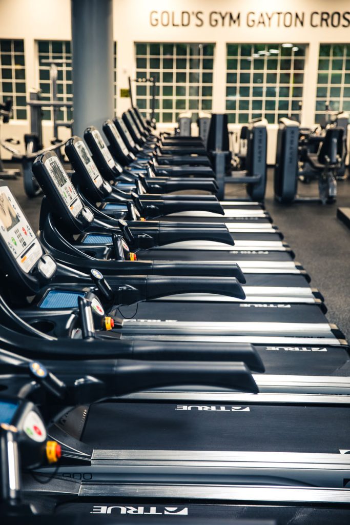 A row of exercise treadmills in a modern gym with large windows in the background and GOLDS GYM GAYTON CROSS written on the wall. The gym appears clean and well-lit, with various equipment visible.