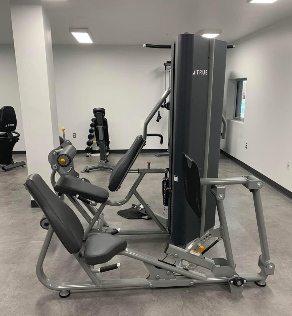 A leg press machine is positioned in the center of a clean, modern gym with gray flooring and white walls. Other gym equipment, including weights and benches, are visible in the background.