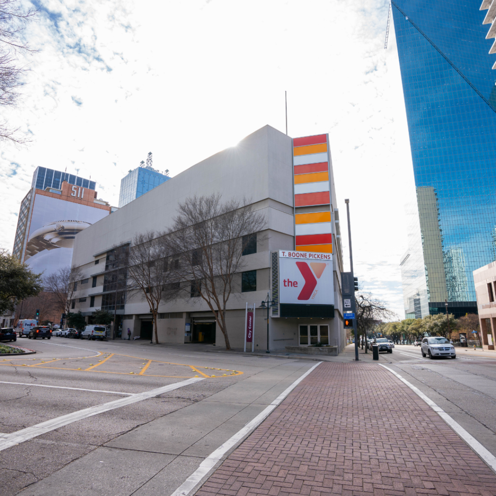 A city street view featuring a multi-story building with a T. Boone Pickens YMCA sign, colorful vertical stripes, leafless trees, and tall glass skyscrapers in the background. Cars are parked along the street.