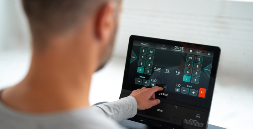 A person operates a touchscreen display on a treadmill, adjusting workout settings. The screen shows options for speed, distance, time, and other exercise metrics.