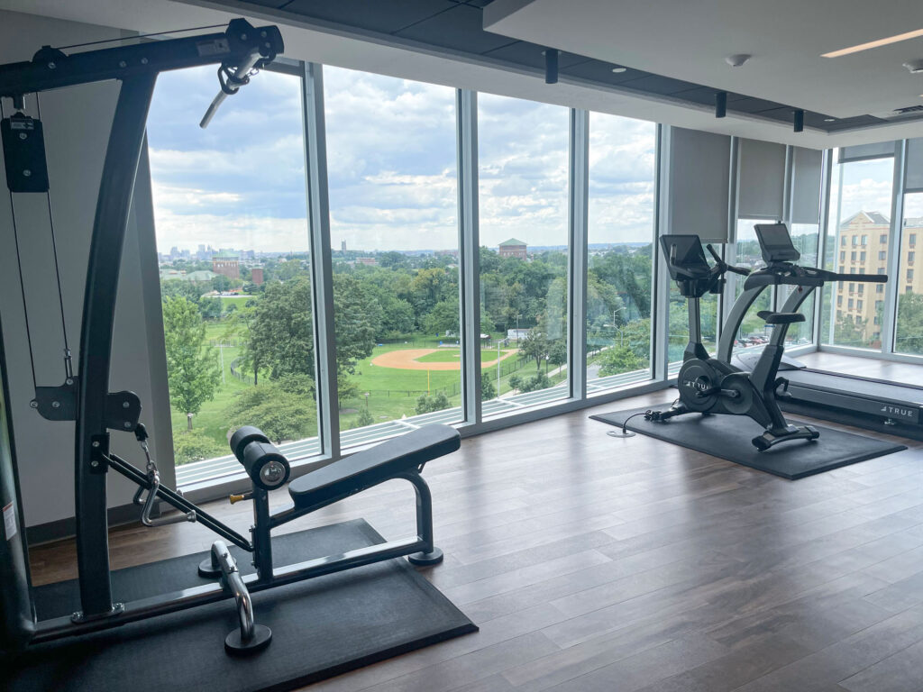 A modern gym with large floor-to-ceiling windows overlooking a green park and baseball field, featuring exercise bikes and weight machines on a wooden floor.