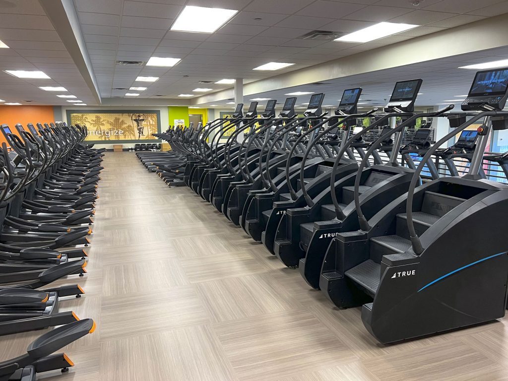 A row of stair climbing machines and treadmills lines the floor of a spacious gym with wood-patterned flooring and bright overhead lights. The wall in the background has motivational text and images.