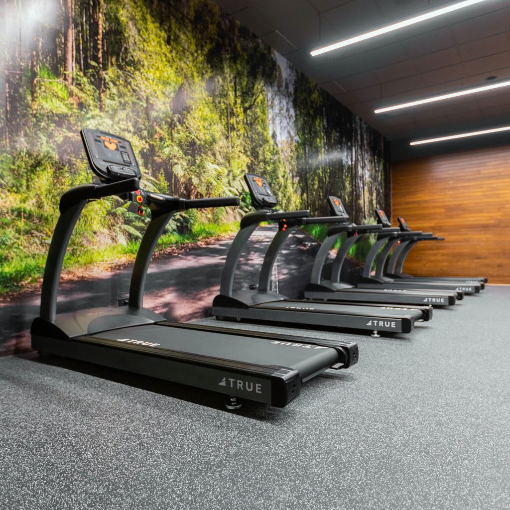Four treadmills are lined up side by side in a gym with a large wall mural of a forest and wood paneling on another wall, creating a nature-inspired workout space.