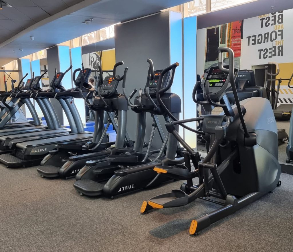 A row of treadmills and elliptical machines in a modern gym with large windows and motivational posters on the walls. The gym is empty and well-lit.