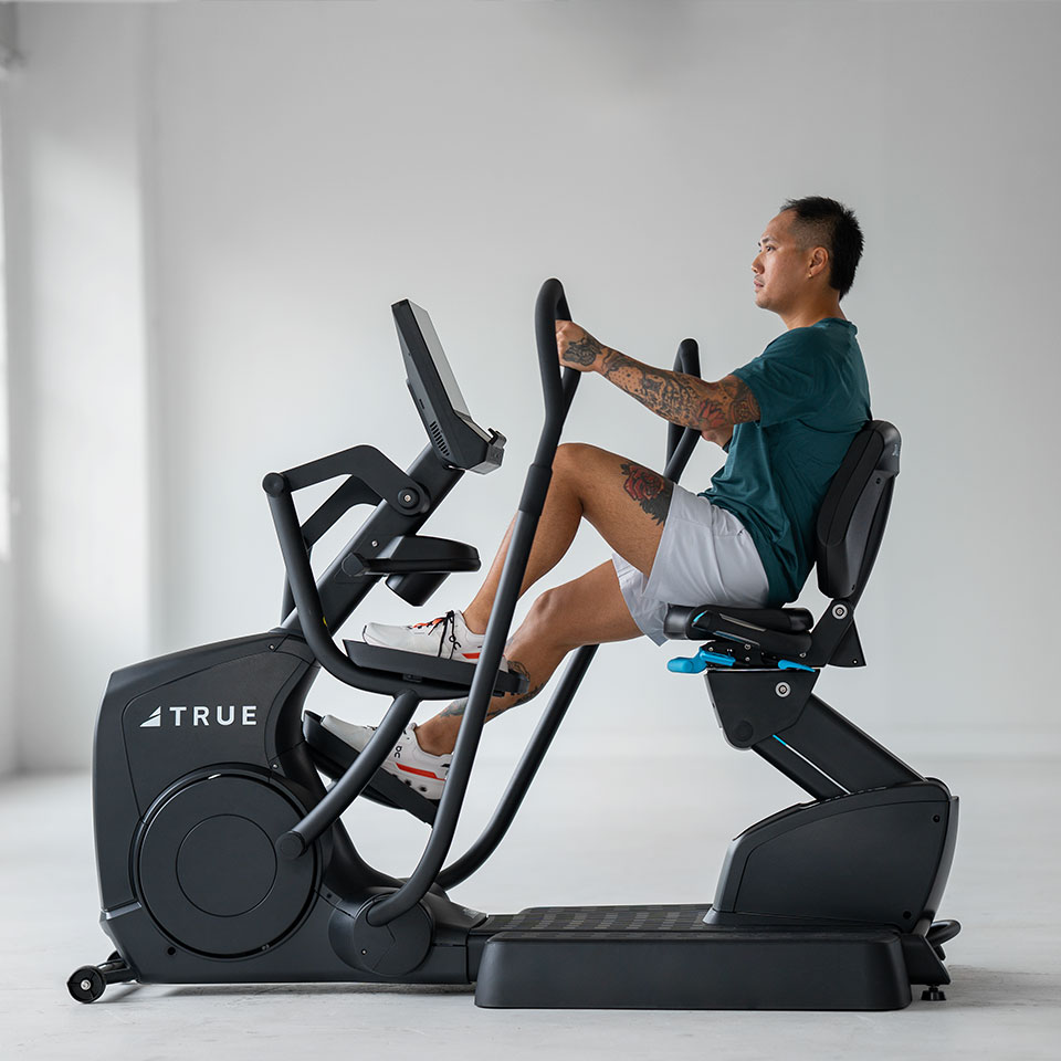 A man with tattoos exercises on a recumbent elliptical machine in a bright, minimal indoor space. He wears a teal shirt, gray shorts, and white sneakers. The machine is labeled TRUE.