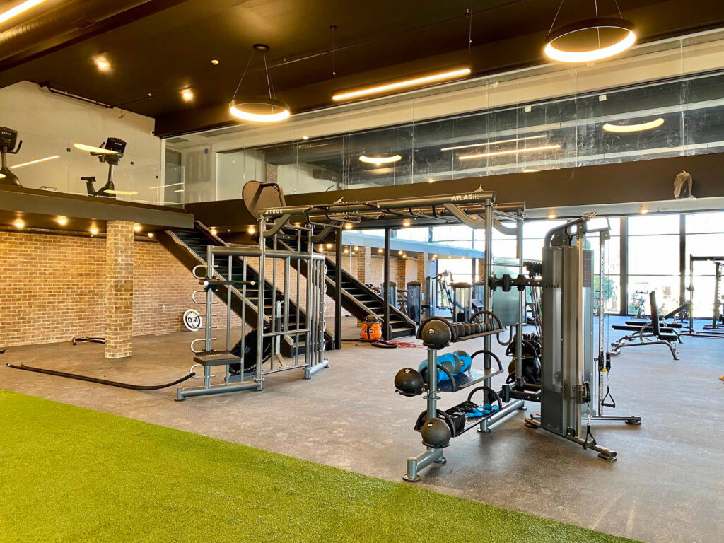 Spacious modern gym with exercise machines, free weights, medicine balls, turf area, and large windows. Exposed brick walls and black ceiling with circular lights add an industrial feel.