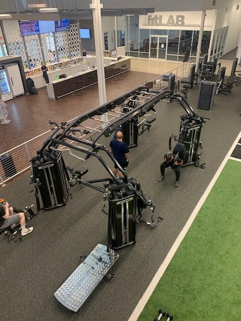 A gym with several people using a large, robotic fitness machine in the center. The area is spacious, with a green turf section, modern equipment, and a glass-walled room labeled fitLAB in the background.