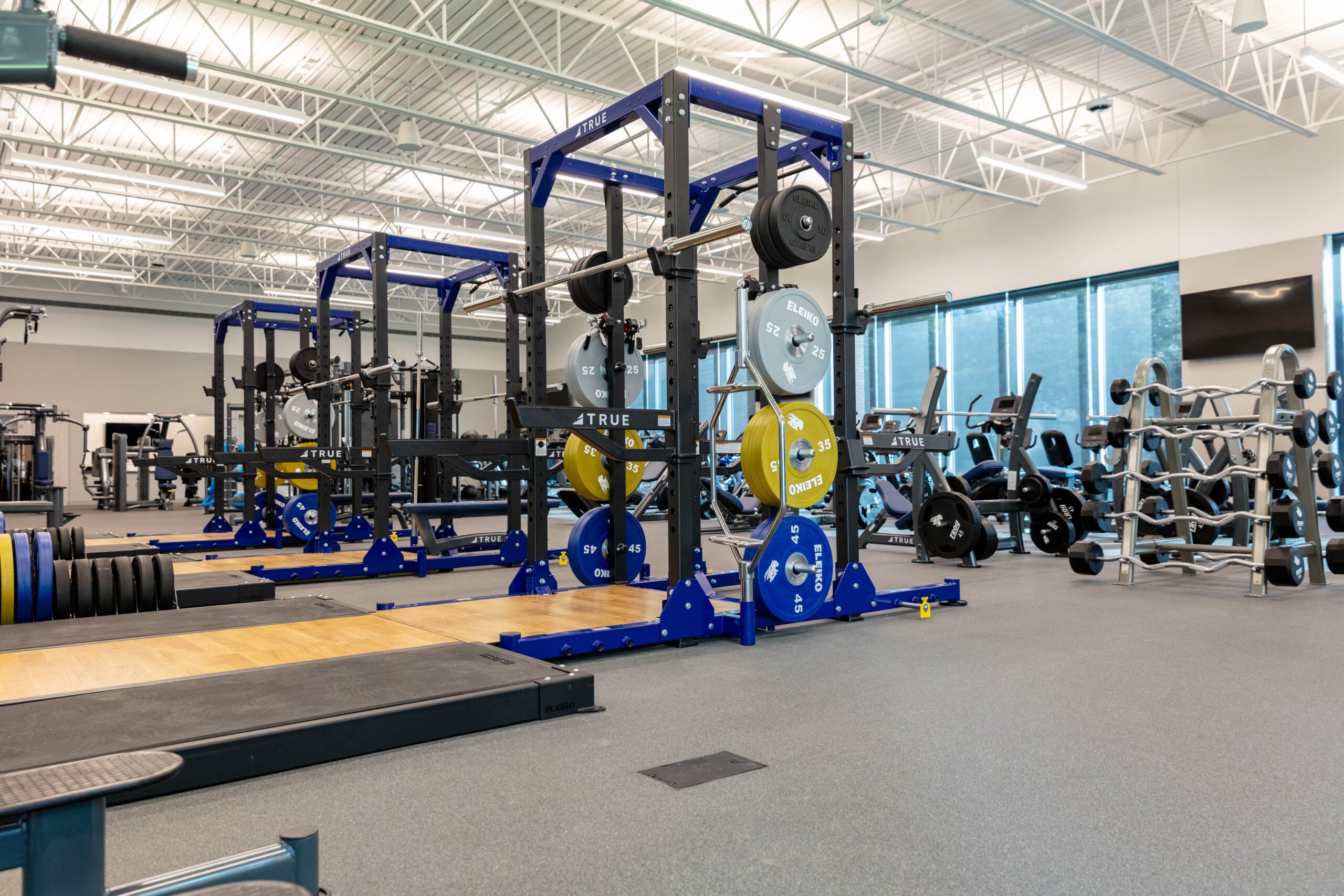 A spacious, well-lit gym with large windows, featuring multiple squat racks with weights, barbells, and various strength training equipment organized neatly throughout the room.
