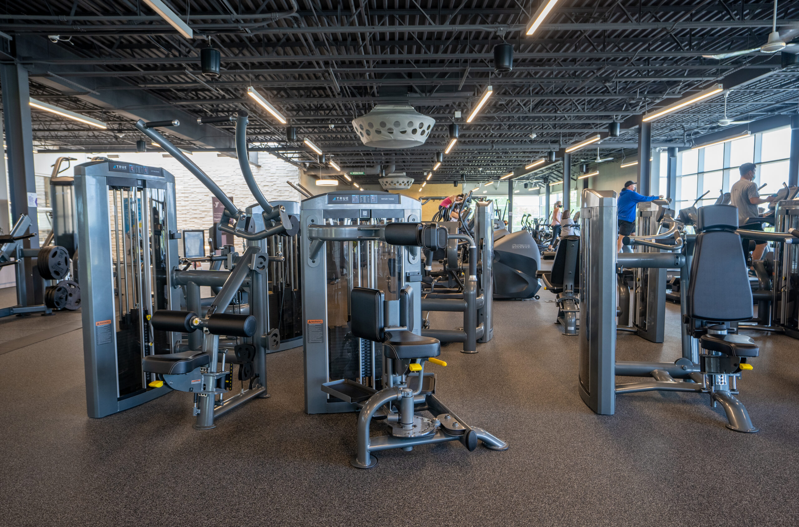 A spacious modern gym with various strength training machines, cardio equipment, and a few people exercising. Large windows on the right let in natural light, and the gym has a high, black open ceiling.