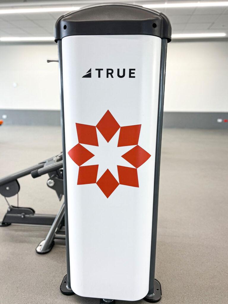 A white fitness machine in a gym has the word TRUE at the top and a large red geometric star design below it. The background is a clean, empty gym with gray flooring and white walls.
