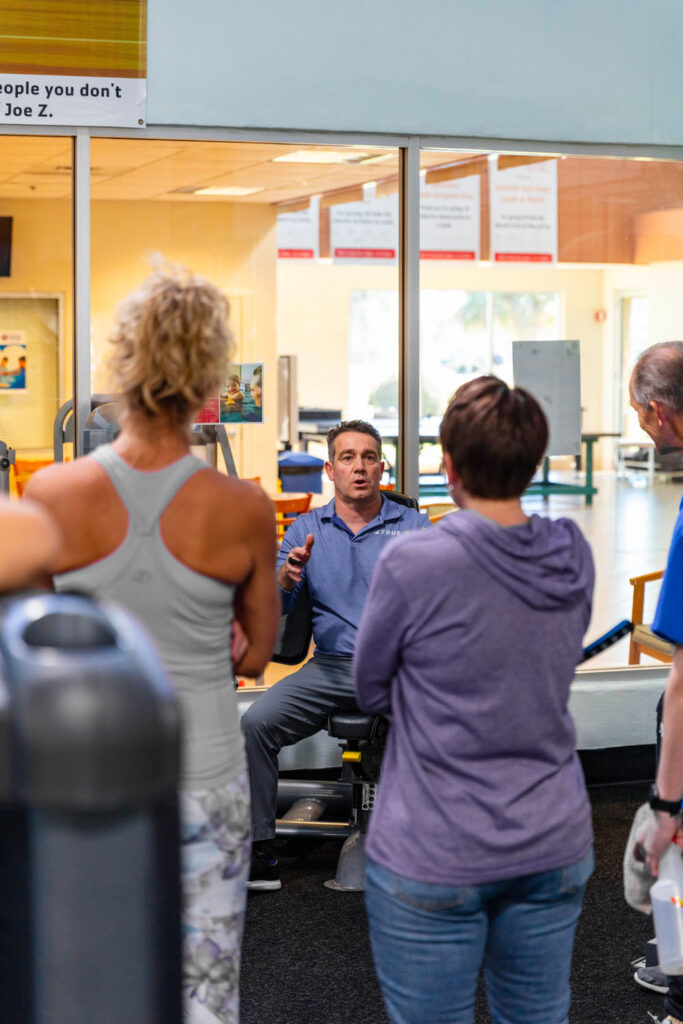 A man in a blue shirt sits and speaks to a small group of adults standing around him in a gym, with exercise equipment and large windows in the background.
