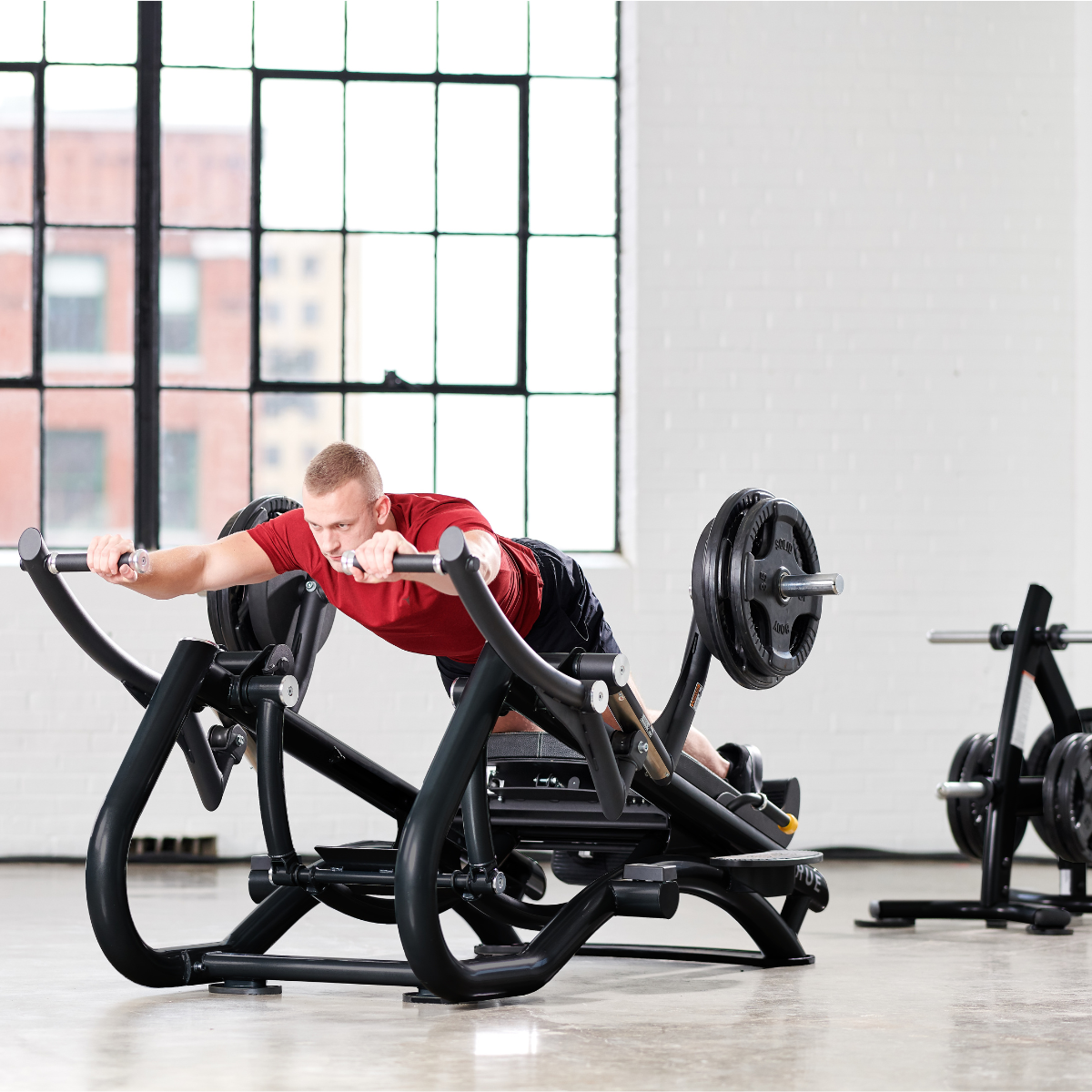 A man in a red shirt uses a weighted prone row machine in a bright gym with large windows and white brick walls. He is gripping the handles and extending his arms while lying face down on the padded bench.