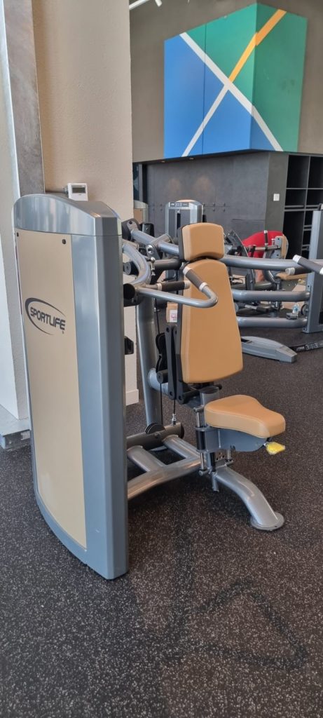 A beige and gray chest press machine labeled SPORTLIFE stands in a gym with black flooring and modern wall art visible in the background.