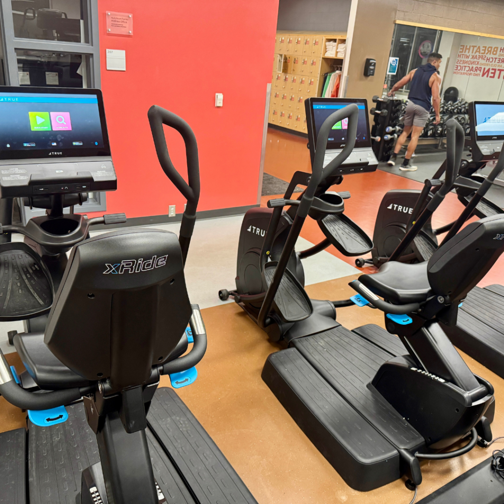 Several stationary exercise bikes and elliptical machines are arranged in a gym with red and brown walls. A person is visible in the background near lockers and rows of dumbbells.