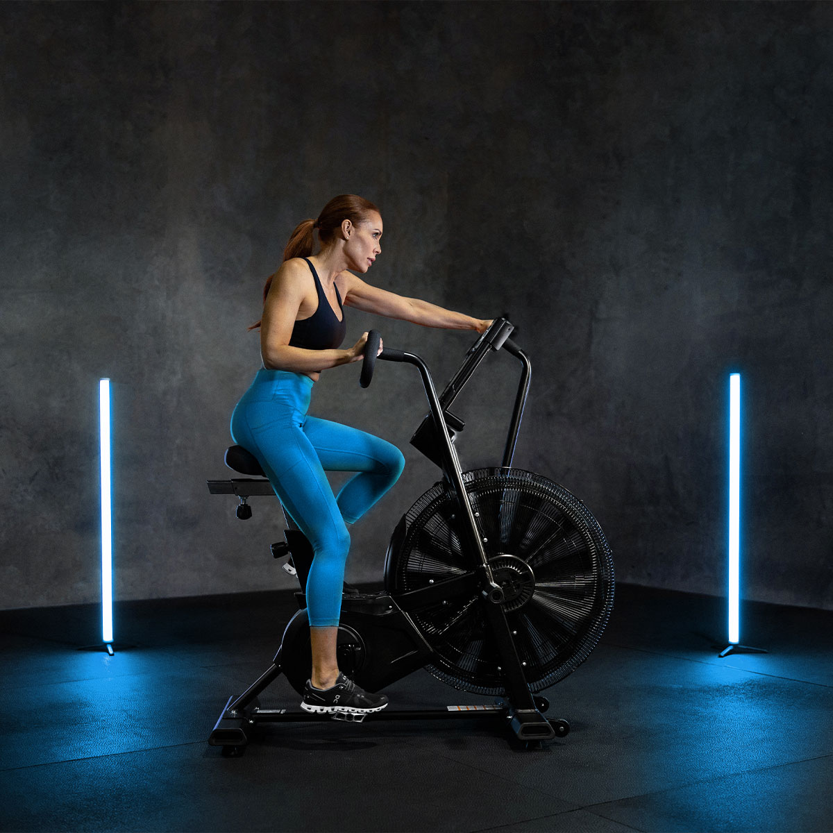 A woman in blue workout leggings and a black tank top exercises on a stationary air bike in a dimly lit gym, with two vertical blue neon lights in the background.