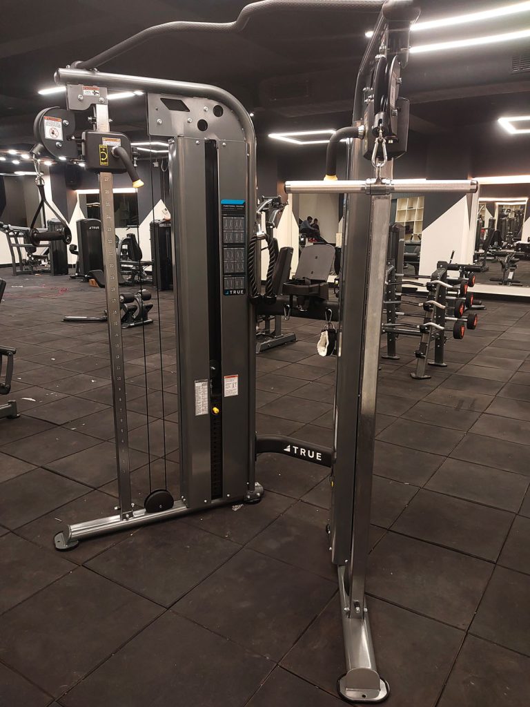 A cable crossover machine with adjustable pulleys and weight stacks is shown in an empty gym, surrounded by other exercise equipment and benches on a black rubber floor.