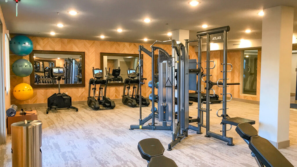 A modern gym with cardio machines, weight equipment, benches, and colorful exercise balls. Large mirrors cover the back wall, and the space is brightly lit with overhead lights.
