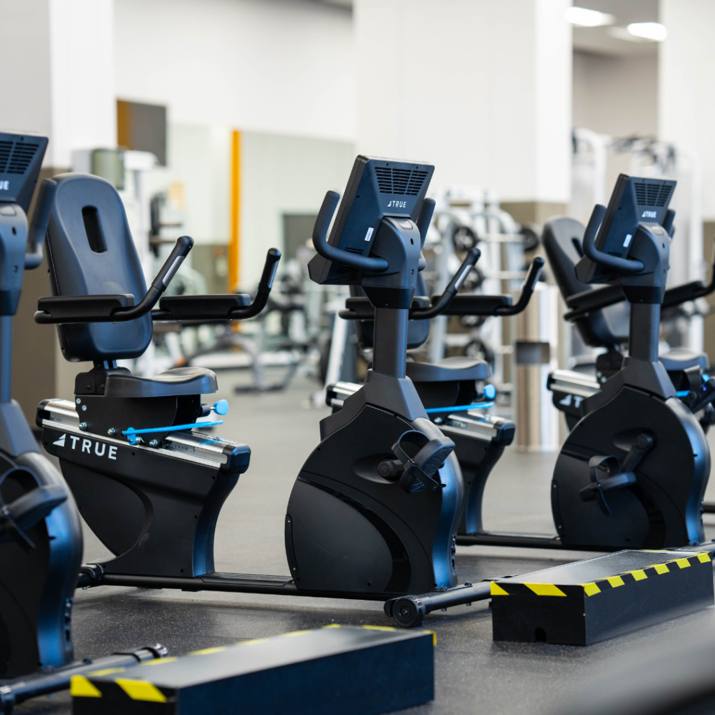 A row of black stationary exercise bikes in a modern gym, positioned side by side on a clean floor, with other gym equipment visible in the background.