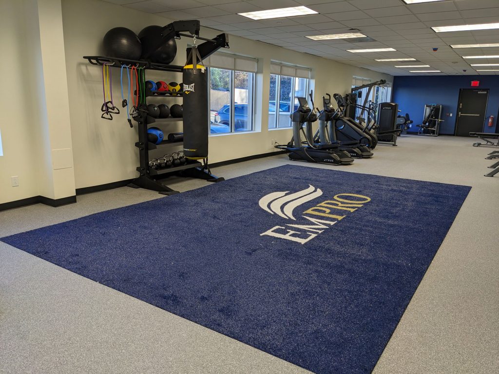 A modern gym with cardio machines, weight racks, resistance bands, and a blue carpet featuring the word EMPIRE with a white and gold logo in the center. Large windows let in natural light.