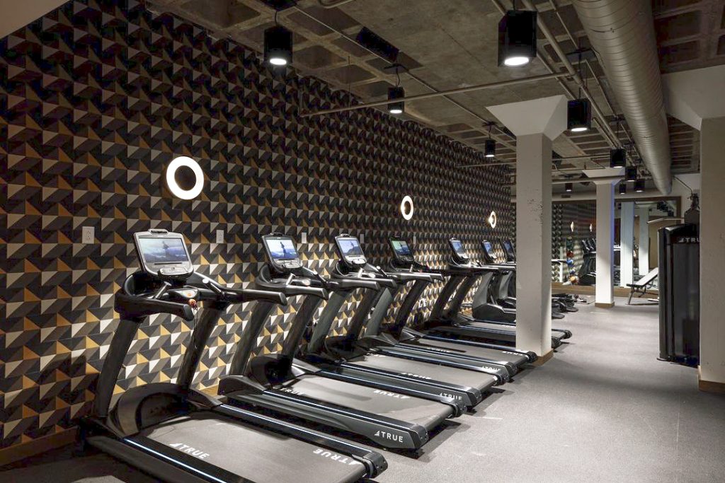 A modern gym with a row of treadmills facing a patterned wall, round lights above each machine, exposed ceiling pipes, and large mirrors in the background.
