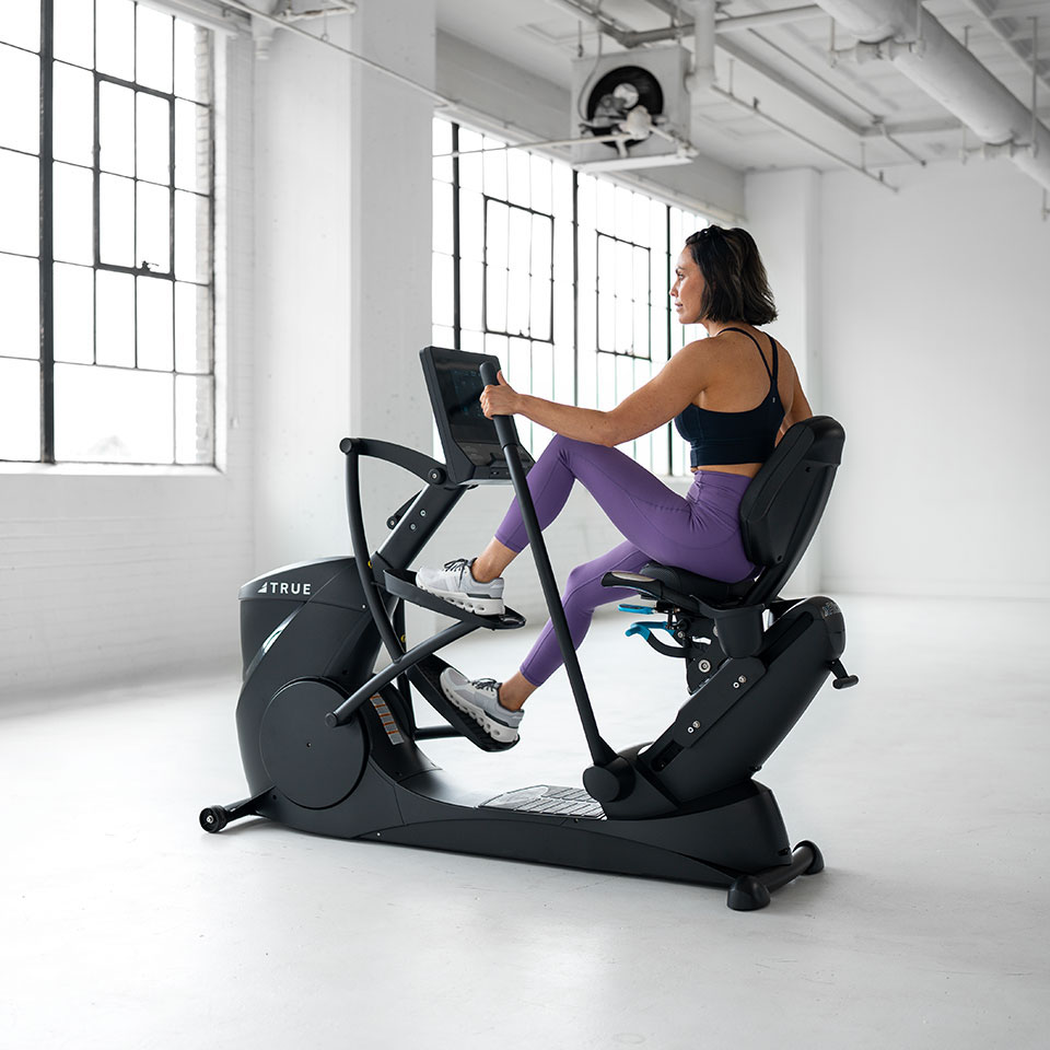A woman in athletic wear exercises on a recumbent elliptical machine in a bright, spacious gym with large windows and white walls.