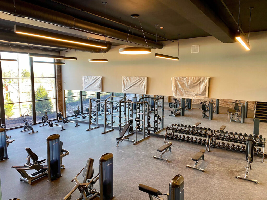 A spacious, modern gym with large mirrors, weight racks, benches, and various strength training machines. Tall windows let in natural light, and the walls are light-colored with minimal decor.