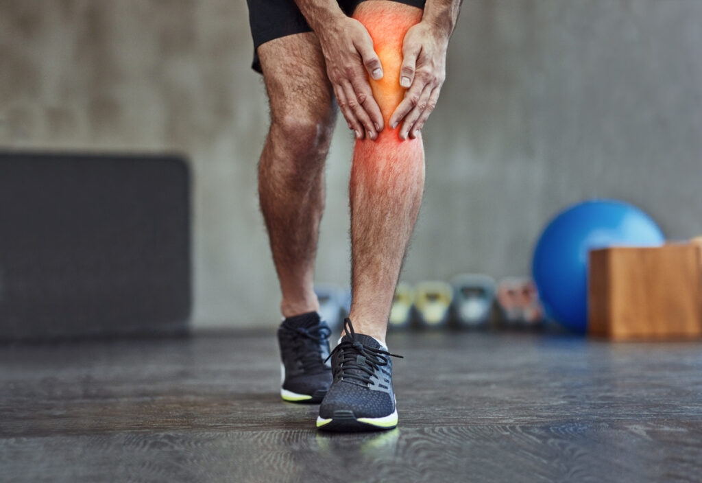 A person in athletic wear holds their knee in pain, with a red area indicating injury or discomfort. The background includes gym equipment such as kettlebells and an exercise ball.