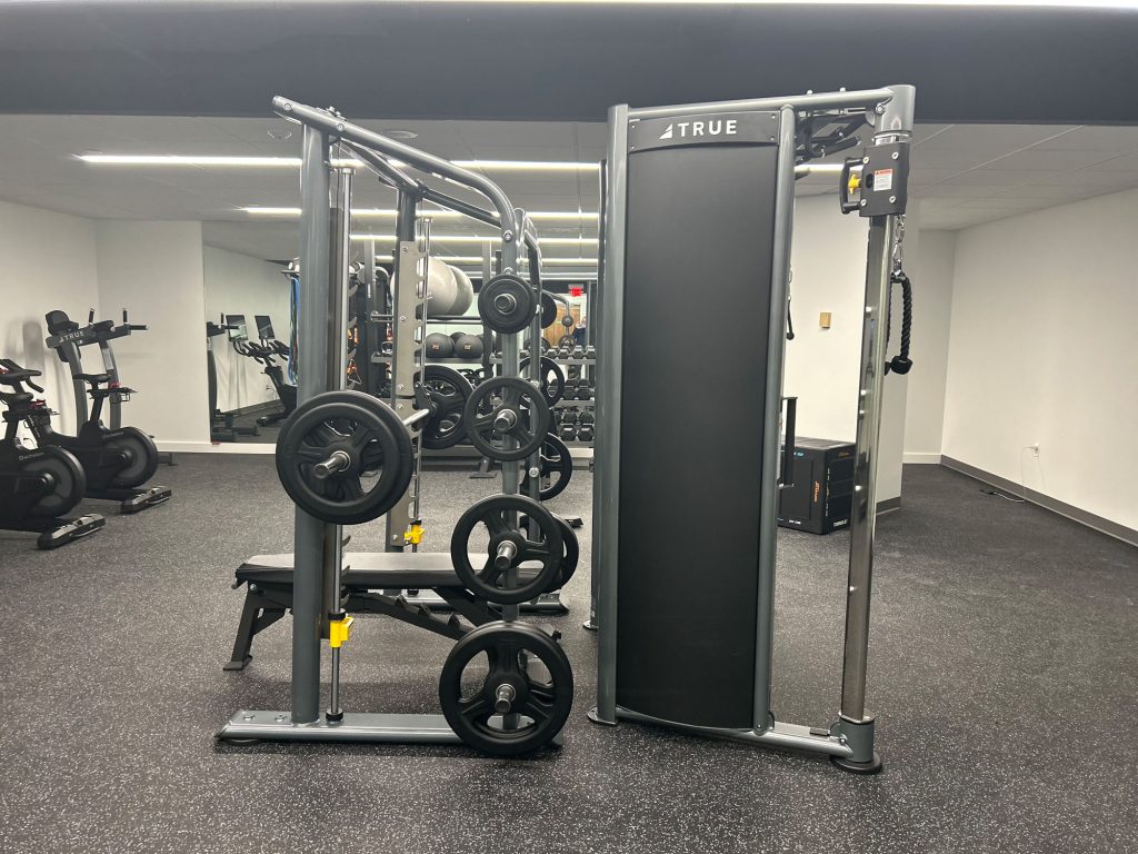 A gym with various exercise equipment, including a weight machine with adjustable plates, a bench, and stationary bikes, set against a mirrored wall in a clean, well-lit room.