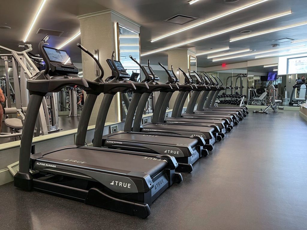 A row of modern treadmills lines one side of a spacious, well-lit gym, with large mirrors on the wall and various exercise equipment visible in the background.