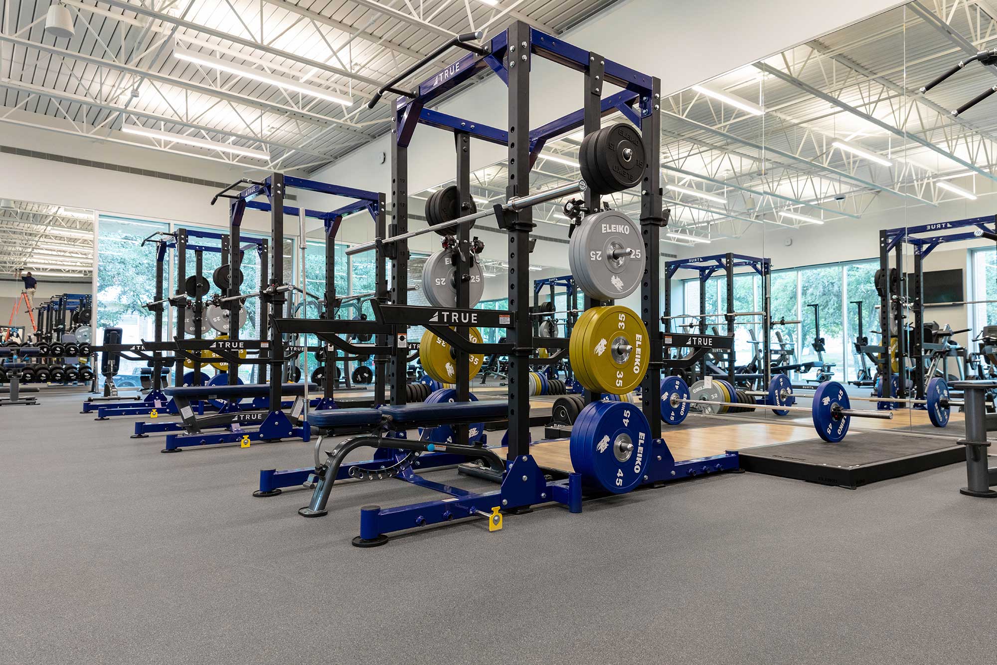 A spacious, modern gym with multiple squat racks, barbells, weight plates, benches, and lifting platforms. Large windows provide natural light, and the area has a clean, organized appearance.