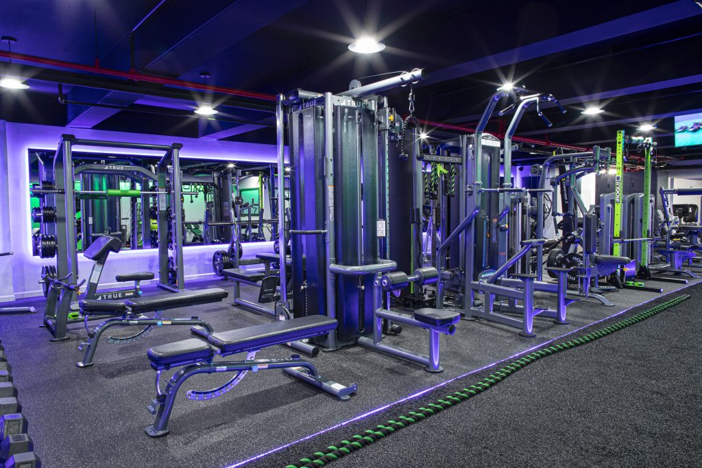 A modern gym with various strength training machines, benches, and free weights. The room is well-lit with blue and purple accent lights, large mirrors, and rubber flooring.