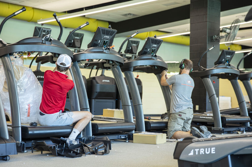 Two workers in casual clothes install or repair treadmills in a gym, surrounded by tools and equipment, with several treadmills lined up in a bright, spacious room.