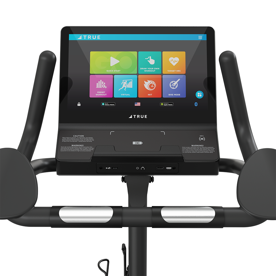 Close-up of a stationary bikes touchscreen display showing workout options and controls. The handlebars and part of the console with caution labels are also visible in the image.