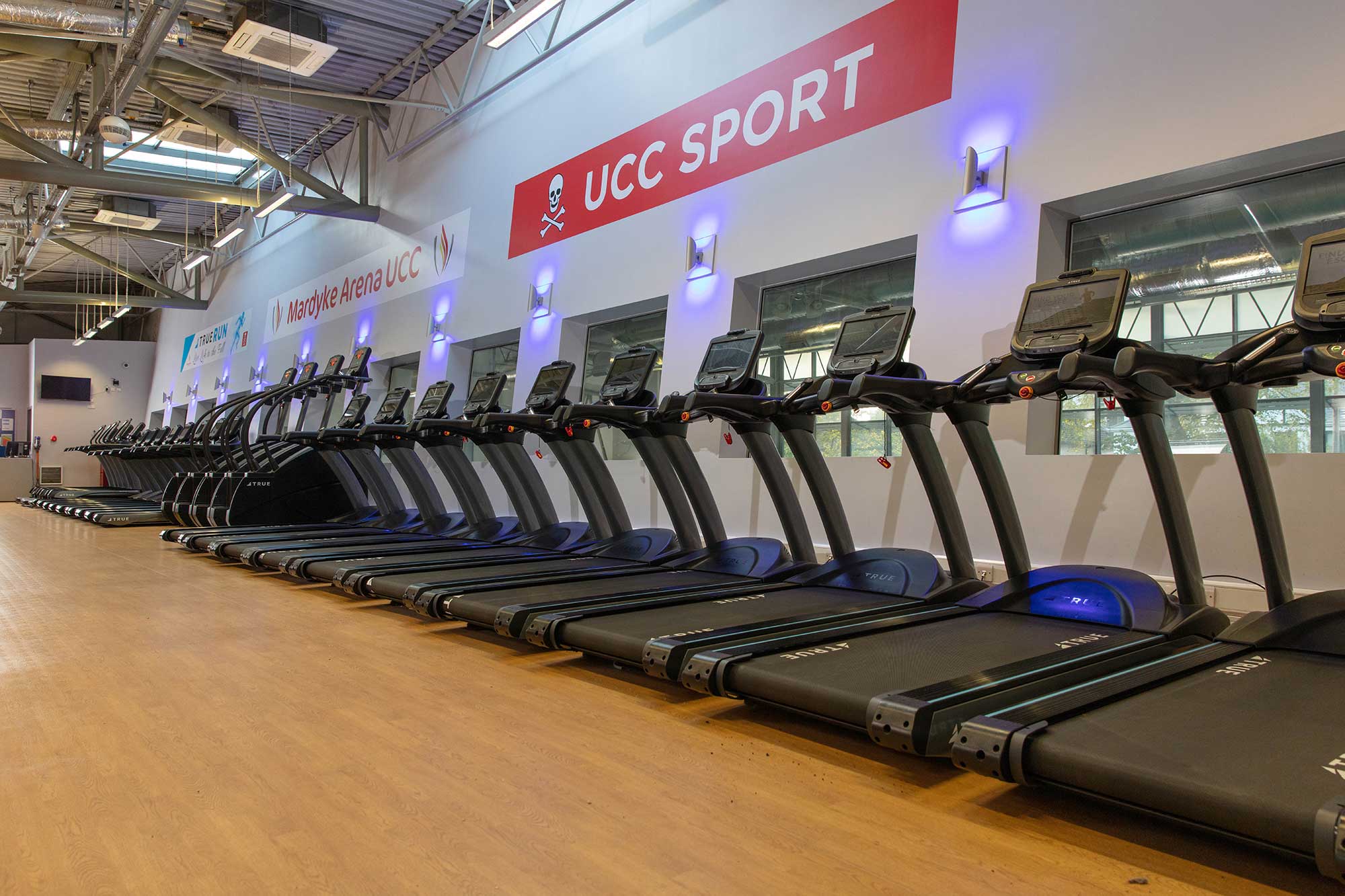 A row of treadmills lines the wooden floor of a modern gym with high ceilings. Large windows and a sign reading UCC Sport are visible on the wall. The gym appears bright and spacious.