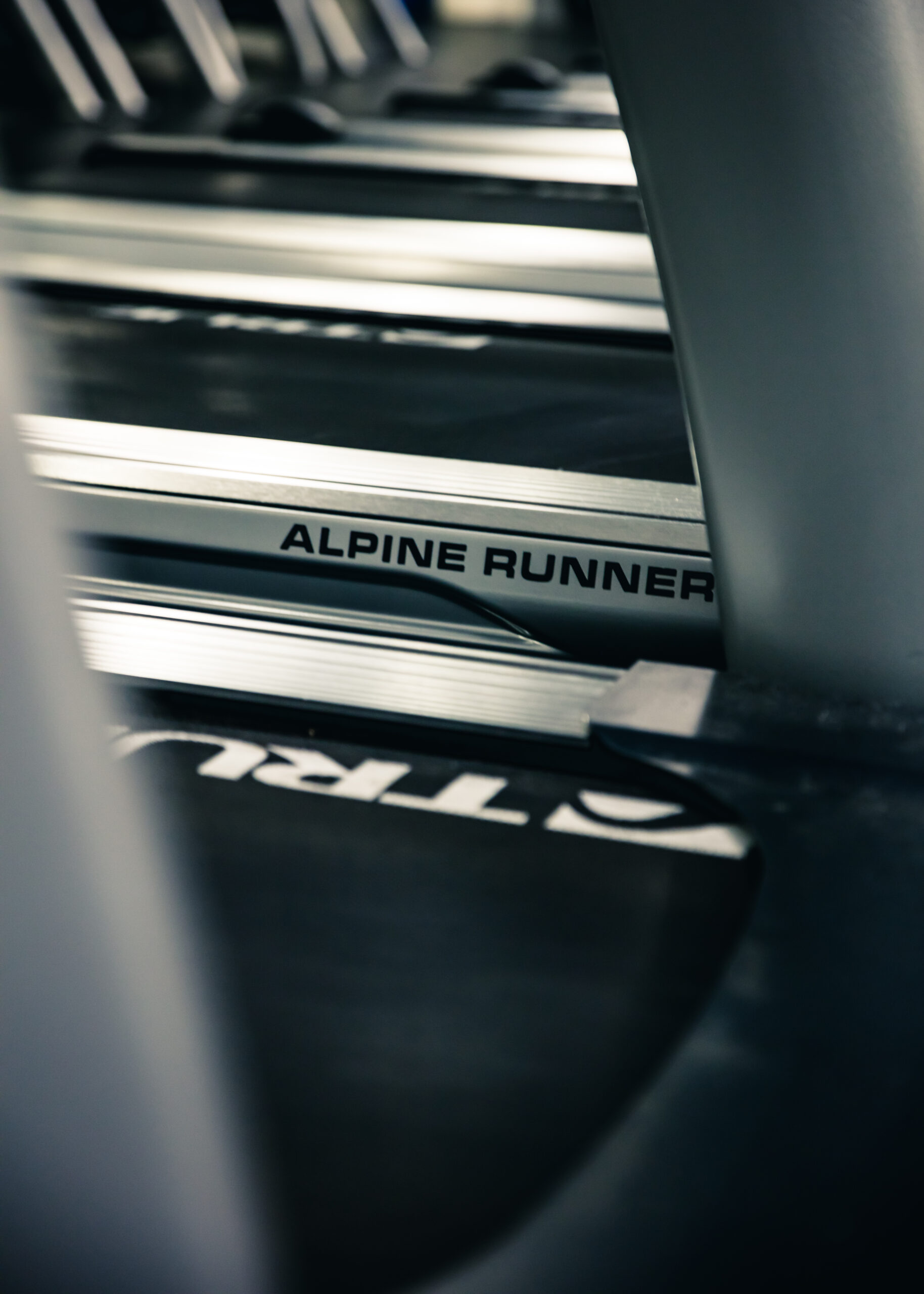 Close-up view of a treadmill with the words ALPINE RUNNER printed on its side, surrounded by other treadmills in a gym setting. The focus is on the treadmill’s frame and text.