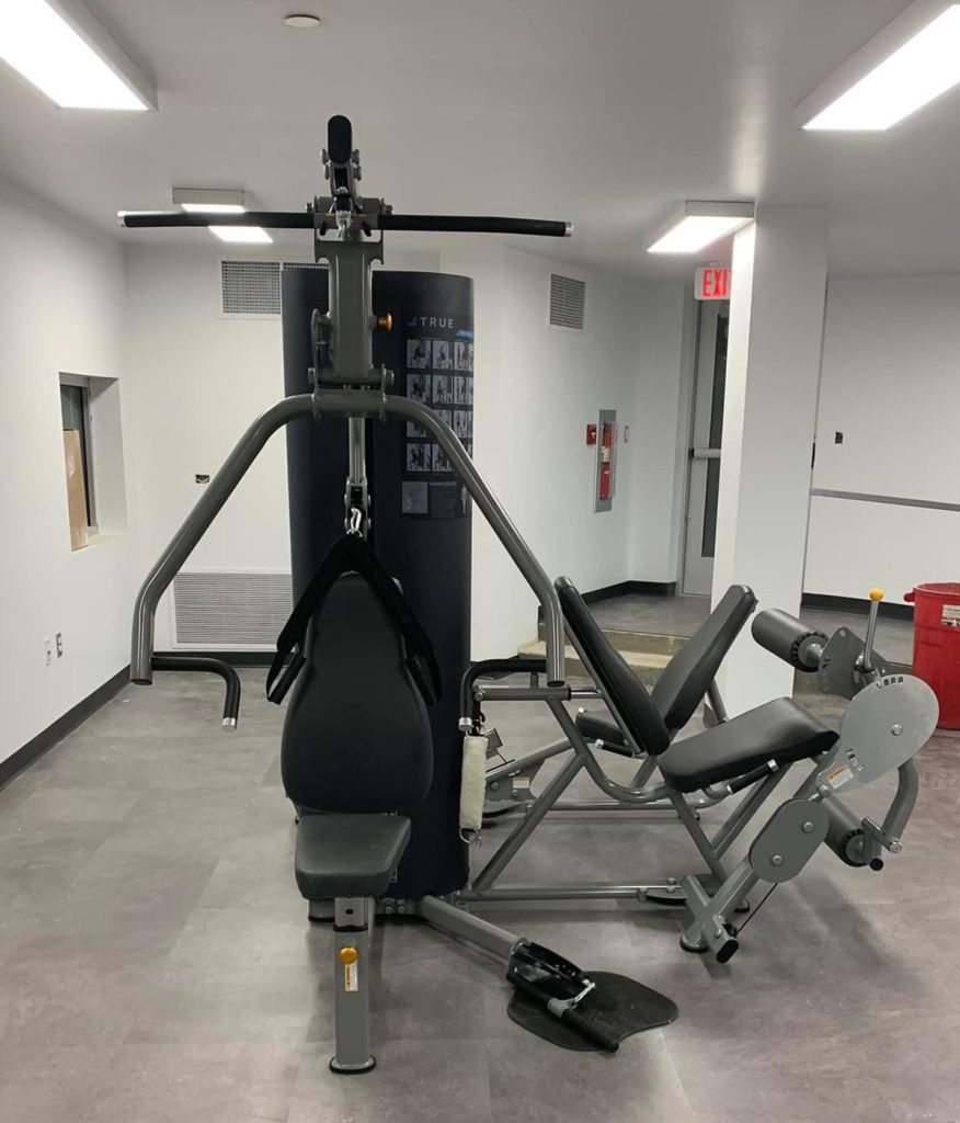 A multi-station gym machine with various attachments for strength training sits in a clean, empty room with gray flooring and white walls. Emergency exit and fire safety equipment are visible in the background.