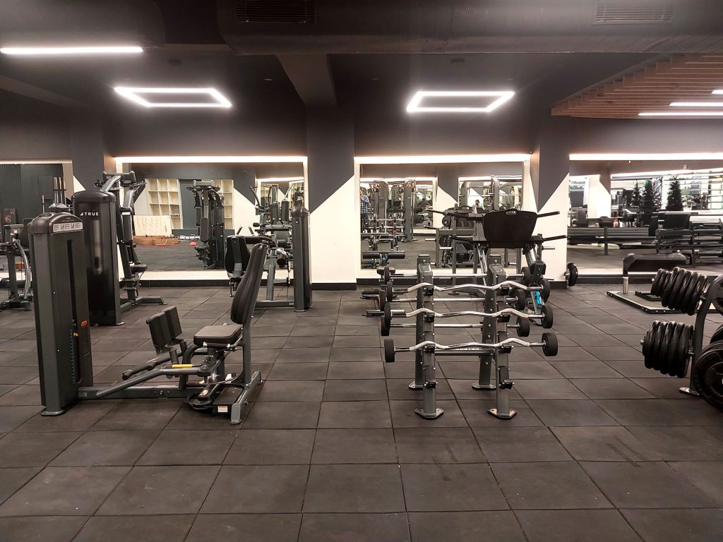 A spacious, modern gym with weight machines, free weights, benches, and large mirrors on the walls. The floor is covered with black mats, and fluorescent lights illuminate the area. The gym appears empty.