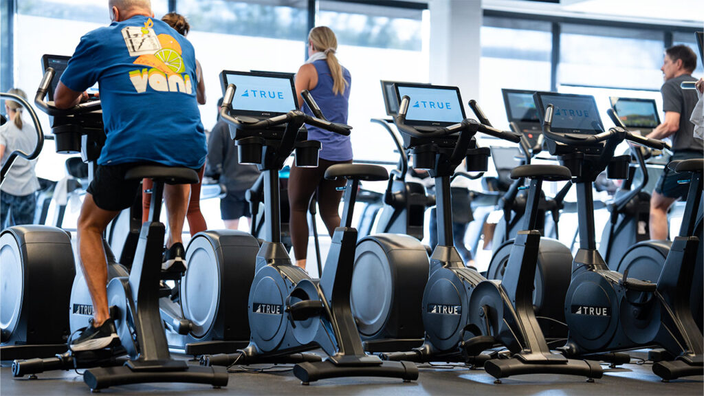 People exercise on stationary bikes and treadmills in a gym. Several exercise bikes are in the foreground, while gym-goers work out on cardio machines in the background. The space appears bright and modern.