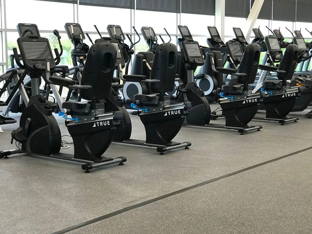 A row of stationary exercise bikes and elliptical machines is lined up in a modern gym with large windows and natural light. The gym floor is covered in speckled grey rubber flooring.