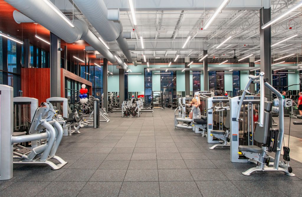 Spacious, modern gym with large windows, various exercise machines, bright lighting, and a few people working out. The floor is covered with rubber mats and the ceiling has exposed pipes and lights.