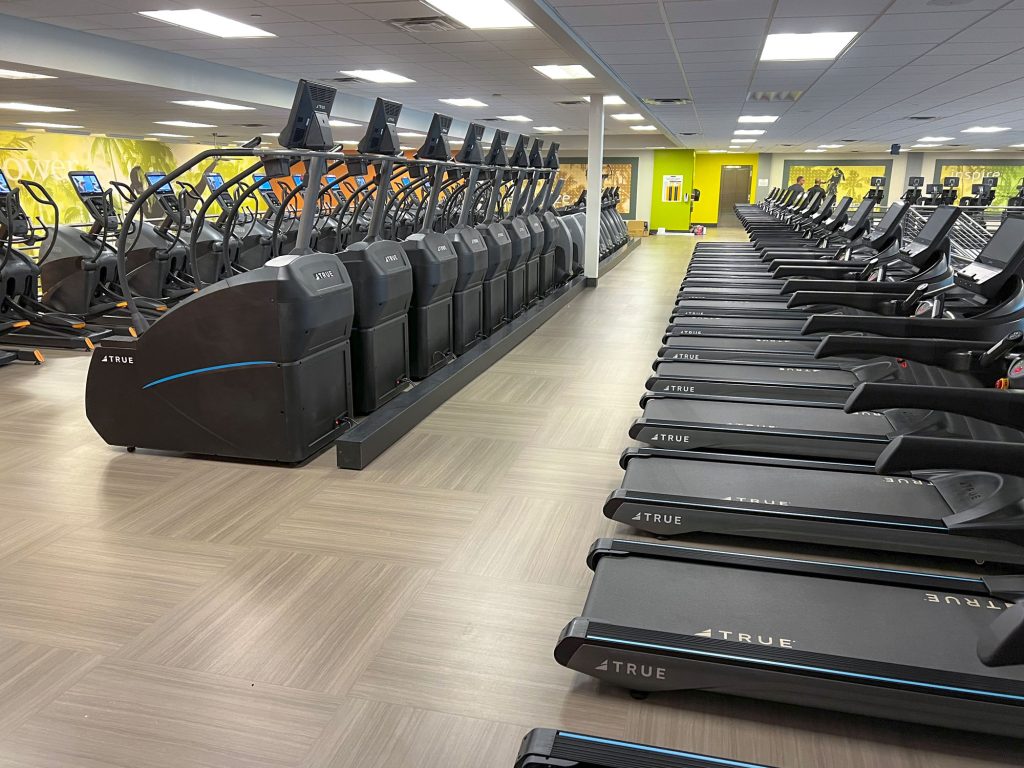 Rows of treadmills and elliptical machines in a spacious, well-lit gym with wood-patterned flooring and bright yellow-green accent walls. The equipment is neatly arranged and the gym appears empty.
