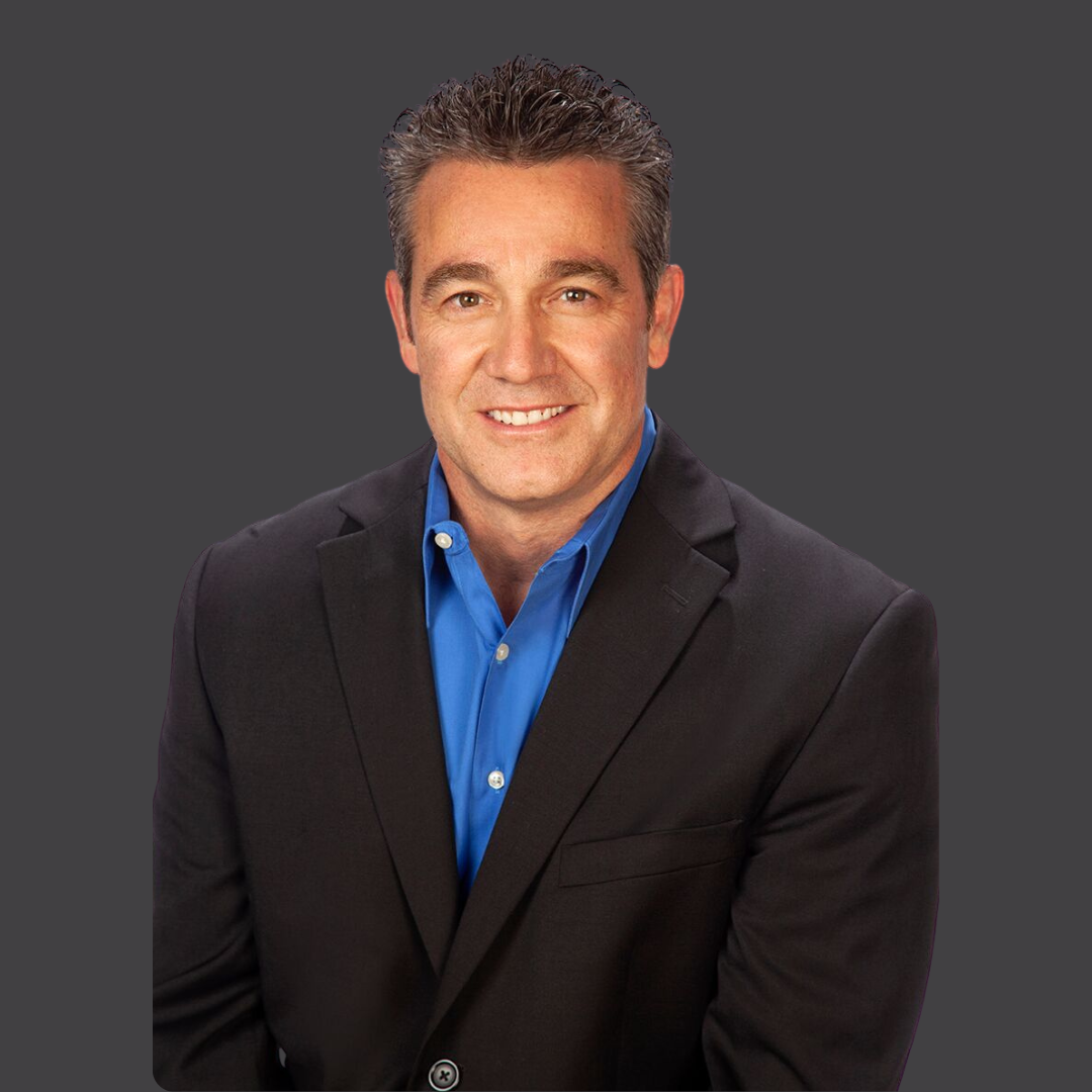 A man with short, dark hair wearing a black suit jacket and blue dress shirt smiles at the camera against a plain dark grey background.
