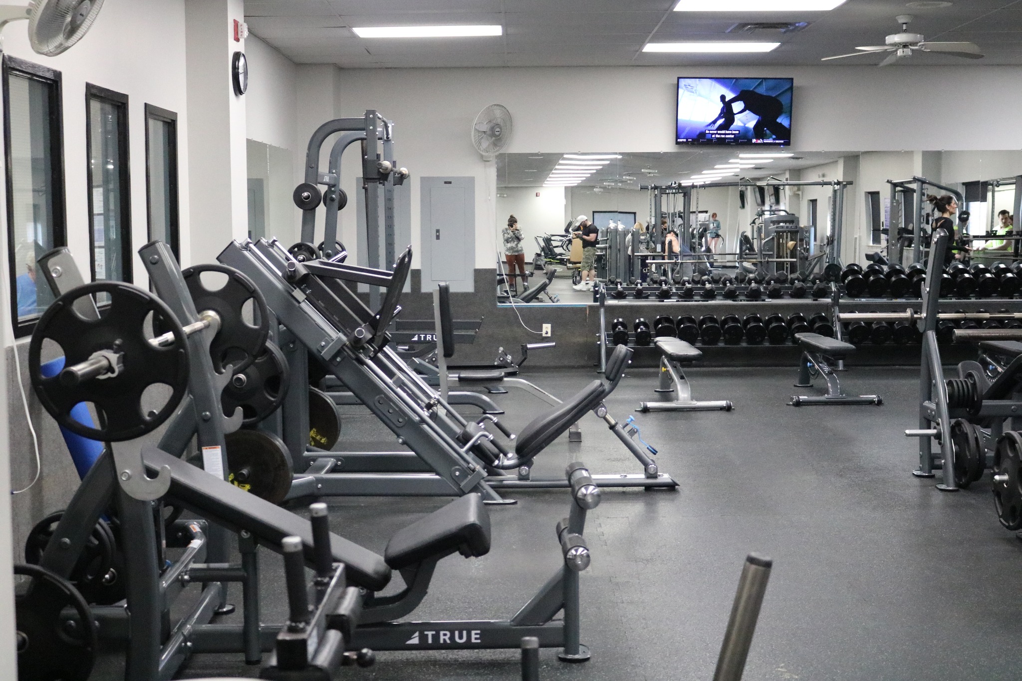 A gym with various weightlifting machines, free weights, benches, and mirrors. A TV is mounted on the wall, and a few people are visible in the background using the equipment. The gym appears clean and well-lit.