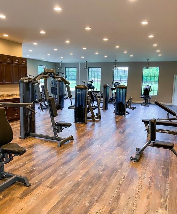 A spacious gym with wooden floors, various exercise machines, and large windows letting in natural light. The room is well-lit, clean, and organized, with equipment arranged neatly throughout the space.