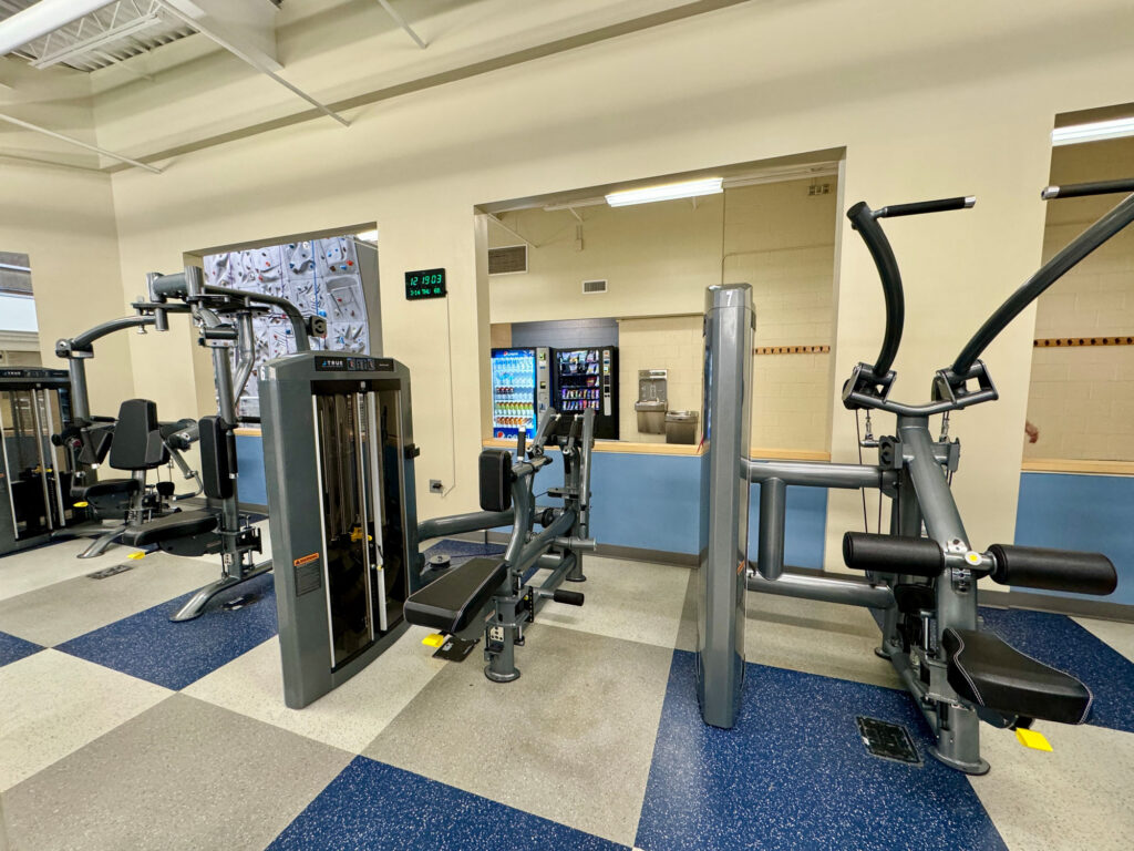 A gym room with various exercise machines, including a leg press and a lat pulldown. A vending machine and water fountain are seen through a window in the back wall. The floor has blue and gray tiles.