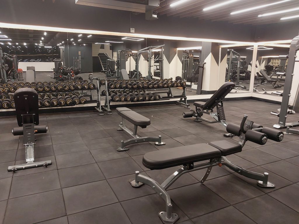 A modern gym with black flooring, benches, weight racks filled with dumbbells, and various strength training equipment. Large mirrors line the walls, reflecting workout machines and the spacious, well-lit interior.