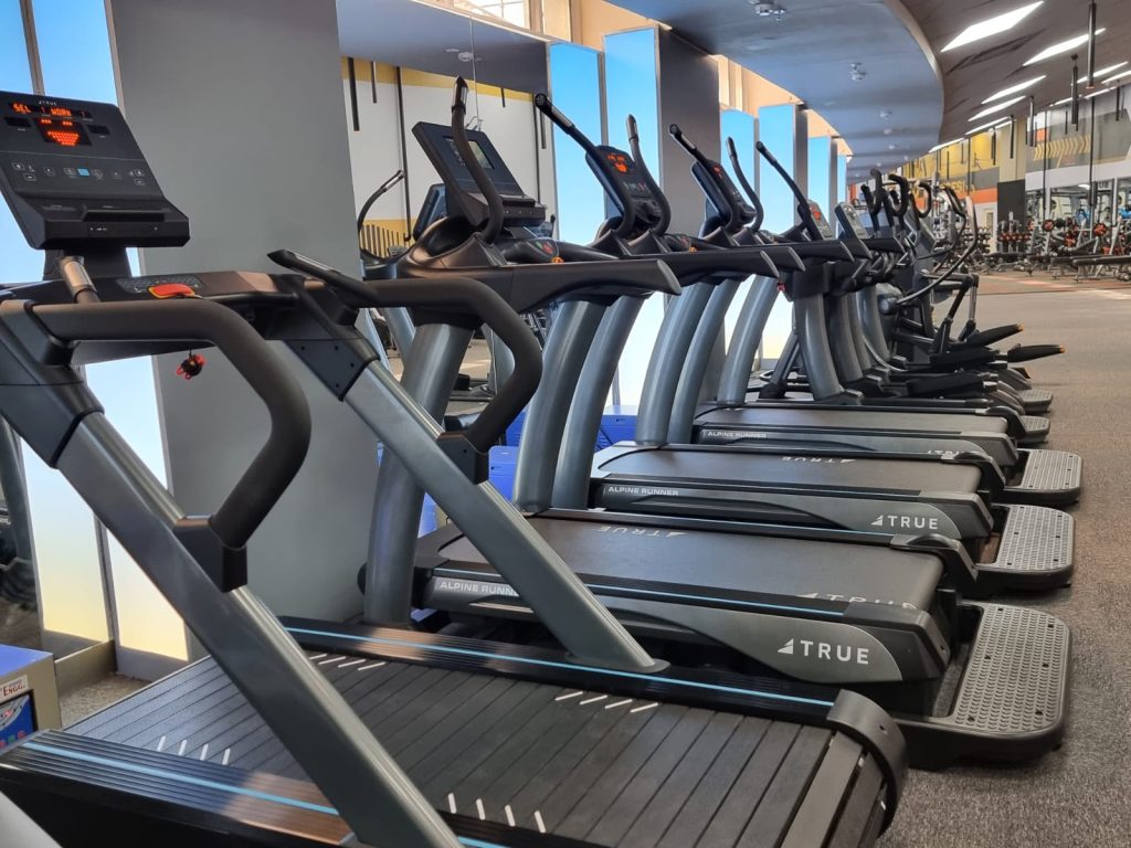 A row of modern treadmills lined up in a well-lit gym, facing large mirrors and windows, with other gym equipment visible in the background.